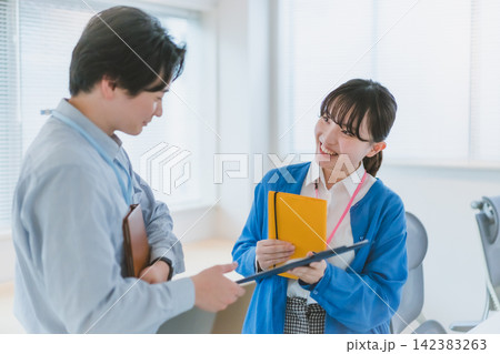 クリップボードを渡しながら笑顔で同僚男性に話しかける女性社員のオフィスラブ 142383263