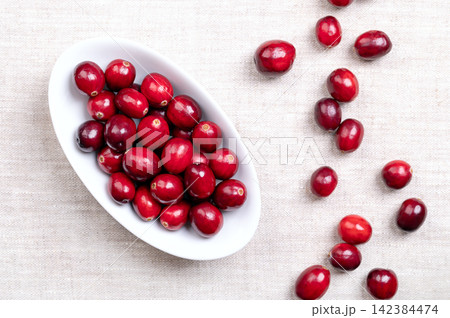 Fresh cranberries, bearberries, or also American cranberries, in an oval white bowl on linen. Ripe, bright red, large cranberries, Vaccinium macrocarpon, with shiny skin and acidic taste. Food photo 142384474