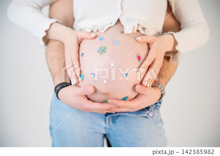 Close-up of a pregnant woman s belly decorated with colorful children s stickers, flowers, hearts and rhinestones, while her husband gently embraces her, celebrating pregnancy, love and the Close-up of a pregnant woman s belly decorated with colorful children s stickers, flowers, hearts and rhinestones, while her husband gently embraces her, celebrating pregnancy, love and the 142385982