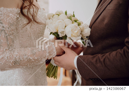Tender close-up of groom holding bride s hands with a beautiful wedding bouquet between them, romantic wedding moment, love, connection and bridal celebration 142386067