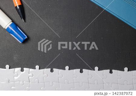 Top view flat lay of office stationery, markers, notebook, and white jigsaw puzzle pieces on a dark chalkboard background with copy space. High Quality Photo Top view flat lay of office stationery, markers, notebook, and white jigsaw puzzle pieces on a dark chalkboard background with copy space. High Quality Photo 142387072