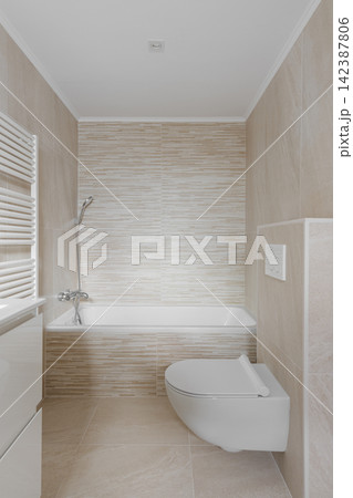 Minimalist modern bathroom interior with bathtub, wall mounted shower, beige textured tile, wall hung toilet, bright neutral finishes, clean residential layout, and refined spa inspired comfort Minimalist modern bathroom interior with bathtub, wall mounted shower, beige textured tile, wall hung toilet, bright neutral finishes, clean residential layout, and refined spa inspired comfort 142387806