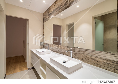 Modern bathroom interior with double countertop sinks wooden accent and large mirror under bright recessed lighting Modern bathroom interior with double countertop sinks wooden accent and large mirror under bright recessed lighting 142387807