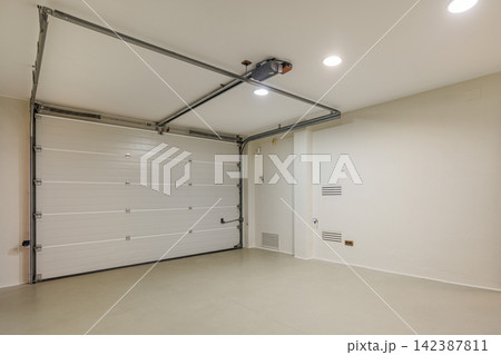 Clean empty residential garage with closed white sectional door bright overhead lighting and smooth light-colored floor Clean empty residential garage with closed white sectional door bright overhead lighting and smooth light-colored floor 142387811