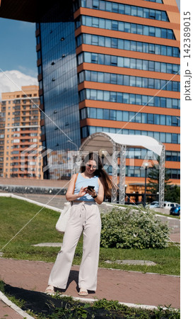 Woman looking at phone, in urban street with modern buildings and traffic light, with woman, phone, white bag, traffic light, with textile, plastic, fabric texture. Woman looking at phone, in urban street with modern buildings and traffic light, with woman, phone, white bag, traffic light, with textile, plastic, fabric texture. 142389015