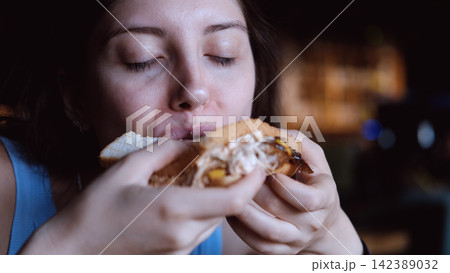 woman eating sandwich in dark setting with contentment close-up hand bread filling food lifestyle blue brown commercial natural light clean copy space editorial educational visual story web. woman eating sandwich in dark setting with contentment close-up hand bread filling food lifestyle blue brown commercial natural light clean copy space editorial educational visual story web. 142389032
