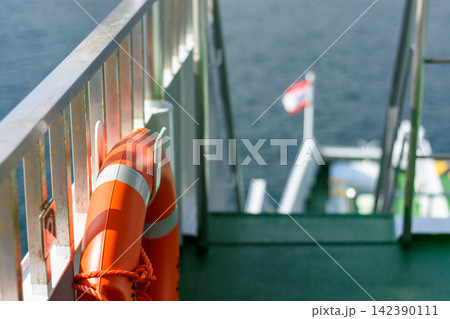 A close-up view from the deck of a ferry showing a bright orange lifebuoy secured with a thick red rope to a metal railing A close-up view from the deck of a ferry showing a bright orange lifebuoy secured with a thick red rope to a metal railing 142390111