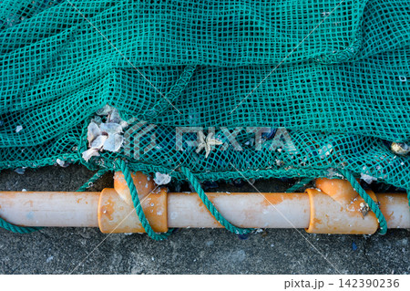 A detailed close-up photograph showing a heavily used green mesh fishing net with accumulated marine debris draped over a cylindrical white pipe and secured with plastic fittings 142390236