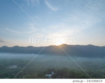 Sunrise emerging behind a forested mountain ridge above misty tropical countryside, with golden morning light illuminating low fog and lush woodland for a peaceful nature. Travel and wellness themes. 142390614
