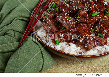 crispy beef, with green onions and sesame seeds, on a rice pad, in a bowl with chopsticks, on a textured surface, no people 142392497