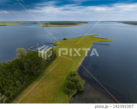 High angle wide horizontal aerial drone photograph of green island spit, small boat marina, Lake Veere, and polder landscape in Ruiterplaat, Zeeland, Netherlands on cloudy summer day 142393307