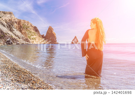 Woman beach ocean standing in shallow water on a pebble beach with majestic coastal cliffs and a sun flare effect 142401698