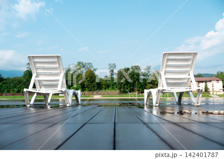 Two white lounge chairs standing on a wet deck with reflections under a blue sky. Peaceful morning view of a poolside terrace, perfect for luxury summer vacation and relaxation concepts. Two white lounge chairs standing on a wet deck with reflections under a blue sky. Peaceful morning view of a poolside terrace, perfect for luxury summer vacation and relaxation concepts. 142401787