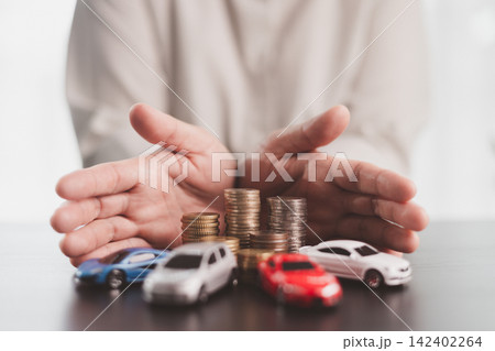 Hands protecting multiple model cars and stacked coins symbolize fleet insurance, vehicle coverage, asset protection, financial security, risk management, and responsible ownership planning. 142402264