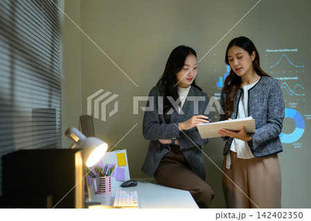 Two Businesswomen Reviewing Data Documents During Office Presentation with Projected Charts 142402350