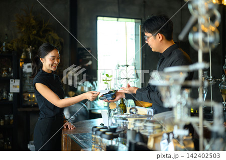 Young Asian Woman Paying with Credit Card at Stylish Boutique Cafe Counter Using Card Terminal Young Asian Woman Paying with Credit Card at Stylish Boutique Cafe Counter Using Card Terminal 142402503