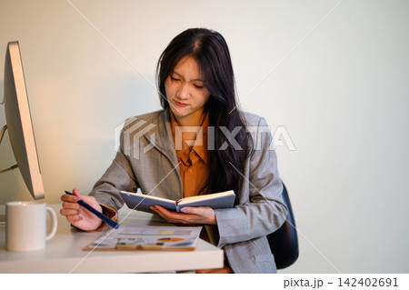 Asian businesswoman reading notebook at office desk with computer monitor and coffee mug, focused and professional Asian businesswoman reading notebook at office desk with computer monitor and coffee mug, focused and professional 142402691