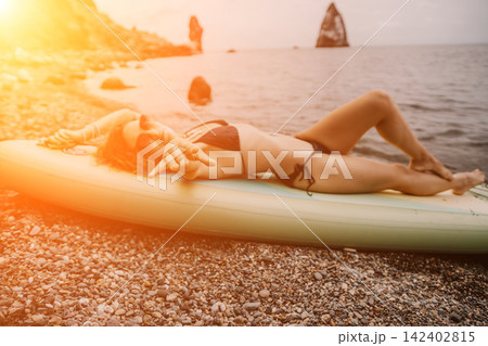 Sunbathing woman, young female relaxing on yellow paddleboard at scenic coastal rocky shore during sunset 142402815