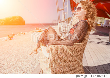 Relaxing vacation atmosphere featuring adult curly woman sitting in wicker chair near sunny ocean coast Relaxing vacation atmosphere featuring adult curly woman sitting in wicker chair near sunny ocean coast 142402818