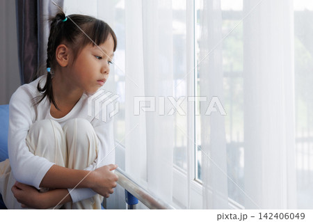 Sad little Asian girl hugging her knees while sitting on hospital bed. Lonely Sick mental health child pediatric patient in hospital room. 142406049