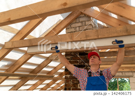 Builder using a spirit level to check the horizontal alignment of new wooden roof rafters during residential construction, ensuring accurate measurements and craftsmanship Builder using a spirit level to check the horizontal alignment of new wooden roof rafters during residential construction, ensuring accurate measurements and craftsmanship 142406175