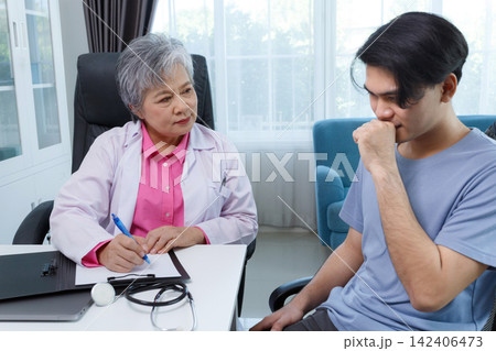 Senior asian female doctor examining and advising young male patient with cough, sore throat, and flu symptoms during medical checkup in clinic office. Hospital treatment and consultation service. Senior asian female doctor examining and advising young male patient with cough, sore throat, and flu symptoms during medical checkup in clinic office. Hospital treatment and consultation service. 142406473