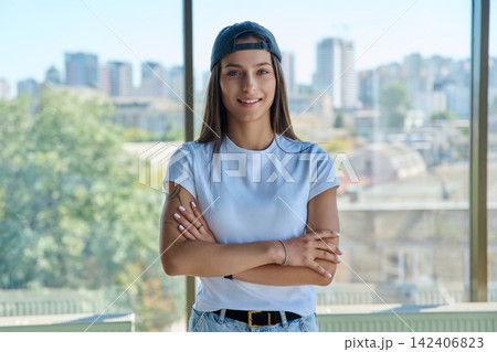 Smiling stylish girl in baseball cap standing by window with city view 142406823
