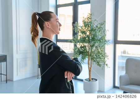 Businesswoman standing profile view crossed arms looking window modern office Businesswoman standing profile view crossed arms looking window modern office 142406826
