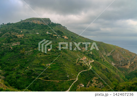 Dramatic overcast sky over lush green mountains with winding trail and distant sea 142407236