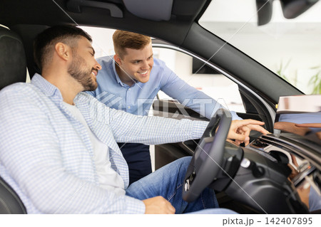 Car sales consultant demonstrating dashboard technology to young male buyer inside vehicle 142407895