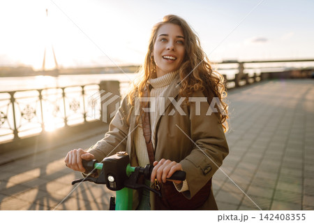 Happy woman driving electric scooter through city. Active life. Ecological transportation concept. Happy woman driving electric scooter through city. Active life. Ecological transportation concept. 142408355