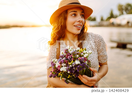 Happy woman in dress sitting on wooden pier near pond and clutched bouquet. Rustic style of wedding. 142408429