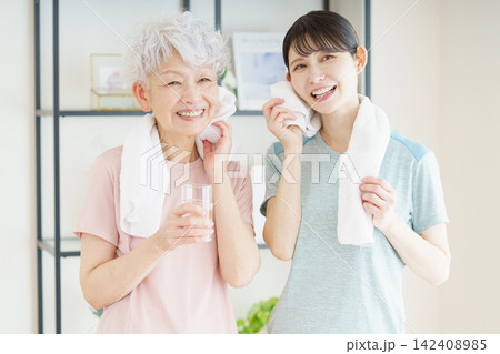 体操の後、水を持ってタオルで汗を拭くシニア日本人女性と見守る若い日本人介護士 体操の後、水を持ってタオルで汗を拭くシニア日本人女性と見守る若い日本人介護士 142408985