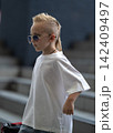 Confident young male child with an edgy blonde mohawk haircut walks down concrete outdoor steps wearing a casual white t-shirt and modern aviator sunglasses. 142409497