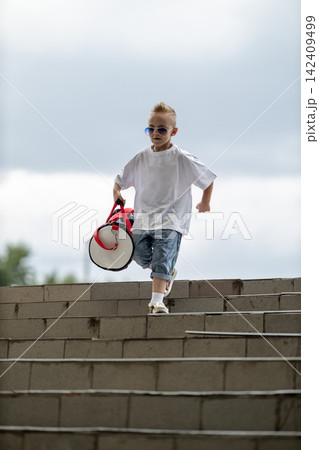 Cool young boy wearing sunglasses and casual clothes descends outdoor concrete stairs while carrying a red and white athletic duffel bag under an overcast sky. Cool young boy wearing sunglasses and casual clothes descends outdoor concrete stairs while carrying a red and white athletic duffel bag under an overcast sky. 142409499
