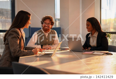 Three diverse colleagues share a relaxed team meeting around a sunlit conference table in a modern coworking office at golden hour Three diverse colleagues share a relaxed team meeting around a sunlit conference table in a modern coworking office at golden hour 142411429