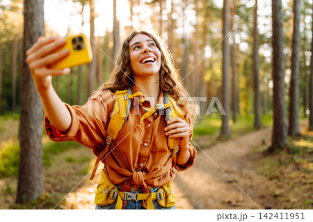 Selfie time! Young woman enjoying a sunny hike in a lush forest with a backpack on a warm day 142411951