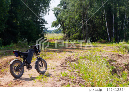 Rugged electric fat bike parked on a dusty dirt trail near a lush green forest, perfect for off-road adventures and eco-friendly outdoor exploration in the summer countryside 142412824