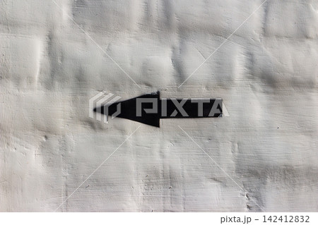 Black spray-painted arrow icon pointing left on a rugged white plastered wall, minimalist urban street art style with a gritty texture and sharp contrast, creative simple navigational symbol 142412832