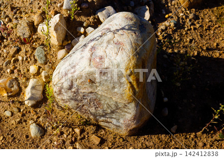 Large smooth quartzite boulder featuring subtle pinkish patterns resting on a sun-drenched dusty ground within a rural sand and gravel quarry under natural daylight conditions 142412838