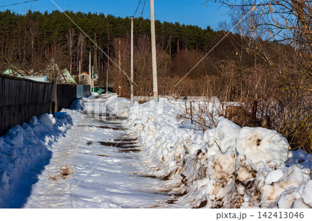 Snow-covered rural path winding between old wooden fences and piles of melting slush under a clear blue sky, capturing the authentic transition from winter to early spring season Snow-covered rural path winding between old wooden fences and piles of melting slush under a clear blue sky, capturing the authentic transition from winter to early spring season 142413046