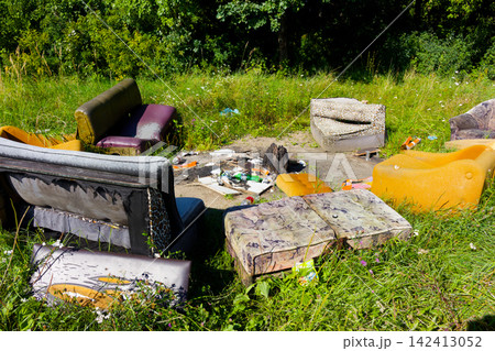 Abandoned mismatched vintage furniture scattered in a grassy meadow forming a derelict outdoor lounge area with litter and debris left behind in a forest clearing 142413052