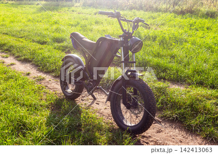 Modern electric bike with fat tires parked on a scenic dirt trail surrounded by lush green grass during a bright sunny day, perfect for eco-friendly outdoor adventure lovers Modern electric bike with fat tires parked on a scenic dirt trail surrounded by lush green grass during a bright sunny day, perfect for eco-friendly outdoor adventure lovers 142413063