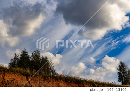 Lush green trees crowning a steep sandy ridge against a dramatic backdrop of swirling wispy clouds and vibrant blue sky on a bright sunny day in nature Lush green trees crowning a steep sandy ridge against a dramatic backdrop of swirling wispy clouds and vibrant blue sky on a bright sunny day in nature 142413103