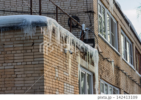 Sharp icicles hang dangerously from the edge of a brick building roof under a layer of frozen winter snow, highlighting the cold urban landscape during a freezing weather season 142413109