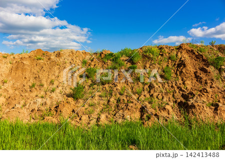 Large pile of raw soil and earth mounds in a green field, showing traces of heavy machinery excavation work under a vibrant blue sky with soft white clouds Large pile of raw soil and earth mounds in a green field, showing traces of heavy machinery excavation work under a vibrant blue sky with soft white clouds 142413488