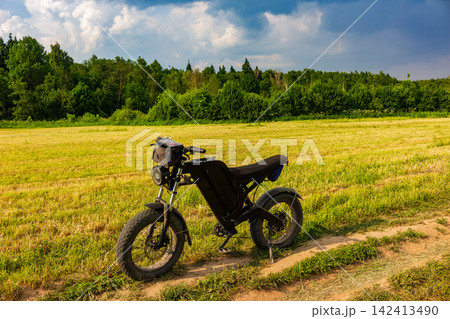 Modern black electric bike parked on a rural dirt path amidst a vibrant summer meadow with a lush green forest background under a bright cloudy sky Modern black electric bike parked on a rural dirt path amidst a vibrant summer meadow with a lush green forest background under a bright cloudy sky 142413490