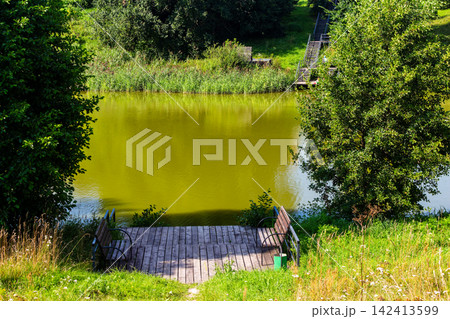Serene countryside pond reflecting lush greenery, featuring a rustic wooden deck with benches as a perfect spot for peaceful relaxation in the warm summer sun, tranquil nature landscape 142413599