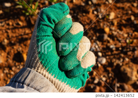 Worker's hand in a green protective glove clenched into a tight fist close-up, symbolizing manual labor, agriculture and gardening against blurry soil background 142413652