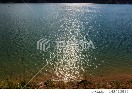 Sparkling blue water surface reflecting bright sunbeams, creating a shimmering effect on the rippling lake expanse, observed from a wild grassy shore during a clear sunny day Sparkling blue water surface reflecting bright sunbeams, creating a shimmering effect on the rippling lake expanse, observed from a wild grassy shore during a clear sunny day 142413985